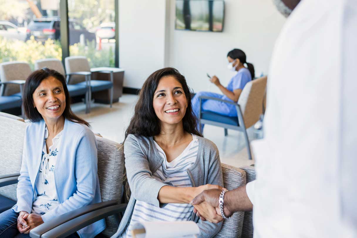 Two women sitting in waiting room, one shaking hand with doctor
